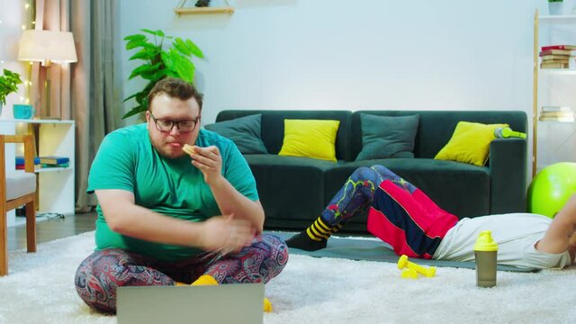 Big fat guy eating a burger while watching a movie on the laptop sit on the floor while other guy doing crunches to get a six pack. Shot on ARRI Alexa