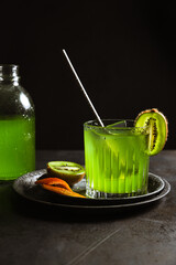 A rocks glass with a refreshing green drink, an ice cube and a bar spoon inside, garnished with kiwi and orange zest. A bottle of drink in the background. Dark backdrop, vertical photo, copy space.