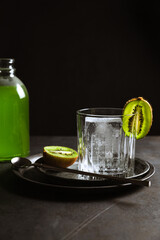 A rocks glass with an ice cube inside, garnished with kiwi, a bottle of green drink, a bar spoon. Dark backdrop, vertical photo, copy space.