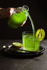 A rocks glass with an ice cube inside, garnished with kiwi, a bar spoon. Pouring a refreshing green drink from a bottle. Dark backdrop, vertical photo.