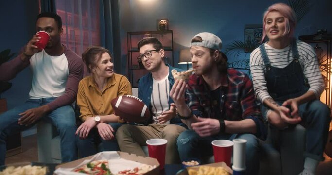 Multi-ethnic happy young male and female friends smiling and speaking while watching sport match on TV at home sitting on couch eating pizza having drinks, soccer fans, entertainment concept
