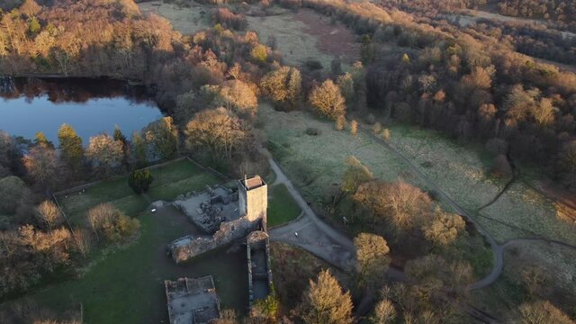 Low-level aerial footage over a small lake and a castle ruin, all surrounded by woodland. 