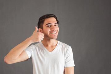 Young smiling funny man in white t-shirt doing happy thumbs up gesture with hand and looking at the camera over gray background, copy space for text