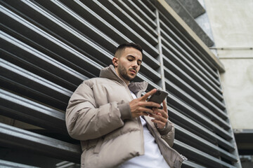 Low angle shot of a stylish European man leaning on a gate and texting on his phone