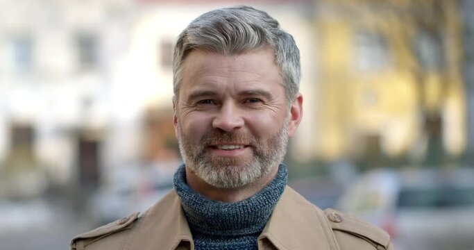 Portrait of Smiling bearded Grey-Haired Man looking into Camera Outdoors. Wearing a trendy Coat and standing on the Street. Attractive Man having healthy Smile. Spending Free Time. Caucasian.