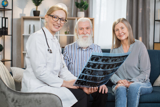 Doctor's home visit. Pleasant positive friendly woman doctor explaining the results of x-ray CT scan of senior bearded man, sitting with his smiling wife on sofa, posing at camera.