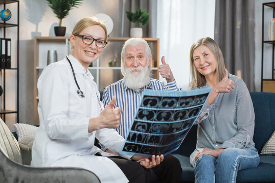 Good news, strong health. Pretty smiling female doctor satisfied of the results of x-ray scan of her patients, happy bearded senior man and pleasant woman , showing thumbs up to camera.