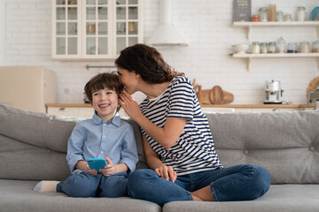 Fototapeta premium Family time at home: beautiful young mother sitting have fun with little son at covid pandemic lockdown, laugh, watch funny video on smartphone and play games. Mum and kid indoors activities concept