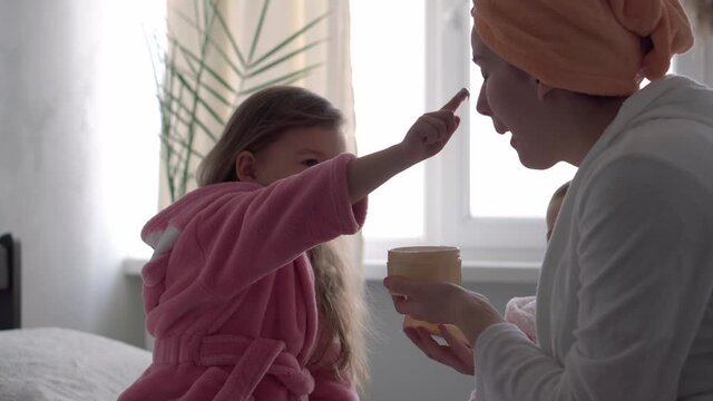 Authentic cute young woman applying face mask of baby girl. mom with 2 children daughters different ages in bathrobes after bath on white bed oposite window. Spa, motherhood, childhood, family concept