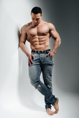 Fototapeta premium A young athlete bodybuilder poses in the studio topless in jeans near the wall. Sport.