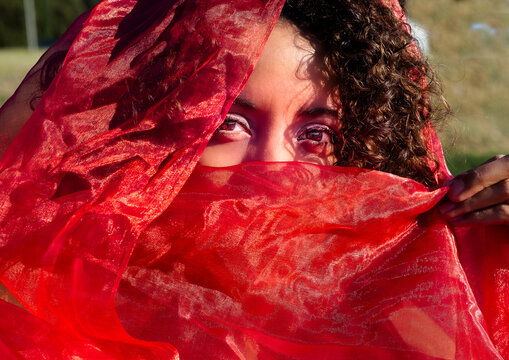 Retrato de mujer joven, morena de cabello corto rizado,  de Honduras, Latinoamericana, mirando a través de de un tul rojo envolviendo su cara en la primavera de 2021.