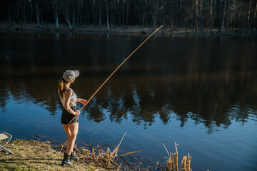 A girl stands on the shore of a forest lake and catches fish with a fishing rod.