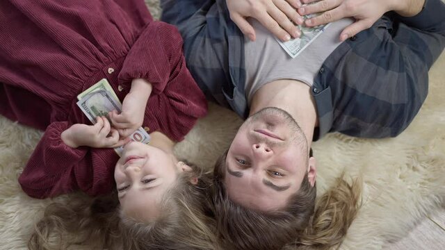 Top View Of Cute Little Girl Sharing Money With Father Lying On Soft Carpet Indoors. Portrait Of Positive Wealthy Caucasian Daughter And Father Resting At Home With Cash. Richness And Family