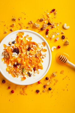 Breakfast concept. Cornflakes, nuts, seeds, berries and honey in a bowl with milk on a bright yellow background. Flakes and nuts scattered on the background. Healthy food. Top view. 