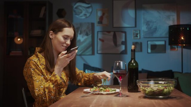 Beautiful woman making voice message on smartphone, having delicious vegetable salad with red wine. Romantic couple dinner online, lovers on long far distance during coronavirus covid 19 lockdown.