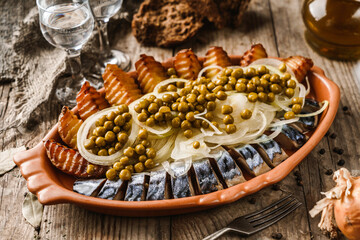 Obraz na płótnie Canvas Fried potatoes with herring fish slices, canned green peas and onions in plate on wooden rustic table with glasses of vodka. Appetizers, close up