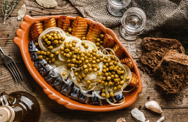 Fried potatoes with herring fish slices, canned green peas and onions in plate on wooden rustic table with glasses of vodka. Appetizers, top view