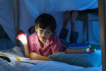 Asian girl child to make a camp to play imaginatively and reading book by flashlight  in living room at home.. - obrazy, fototapety, plakaty