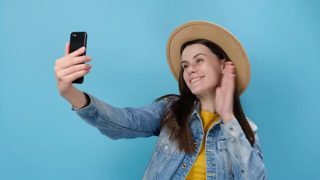 Cheerful European young girl doing selfie shot on smartphone blowing send air kiss put hand on head cheek, dressed in denim jacket and hat, isolated on blue studio background. People lifestyle concept
