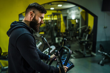 Fototapeta na wymiar Adult caucasian male athlete training on the treadmill at the gym - back view of man with beard in black hoodie running on the treadmill sport and fitness concept back view copy space