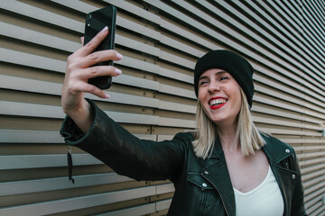 Young woman, taking a selfie with her cell phone. Smiling. With a wall of the street in the background. Women, smartphones and lifestyle concept.
