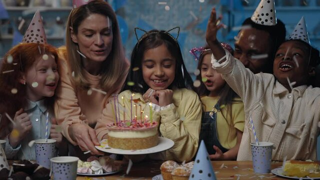 Cute kid with black hair waiting with closed eyes while woman bringing and putting birtday cake in front of her. Girl blowing candles on cake while having party and confetti falling on her.