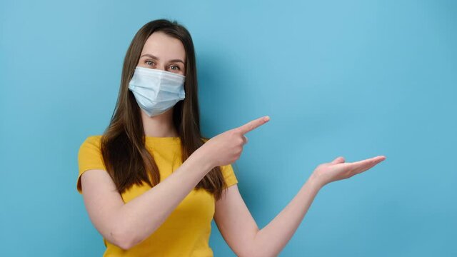 Young Caucasian woman in medical protective face mask keeps raised palm, points with forefinger at free space, pretends holding cool product, wears yellow t-shirt, isolated over blue studio background