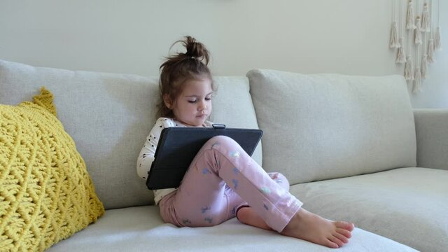 Cute little kid girl using digital tablet technology device lying on sofa alone. Small child hold computer surfing Internet play game at home. Children tech addiction concept.