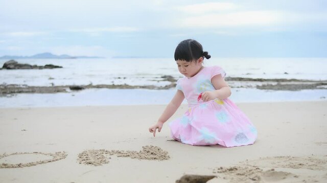 Playful little Asian girl kid playing with sand on a beach, having fun using finger writing on sand ocean beach, creative imagination creativity of childhood thinking, on summer holiday vacation