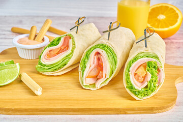 Light breakfast of three ham and cheese wraps on wooden board with healthy orange juice