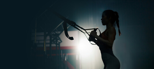 Silhouette of young Sexy fit Asian woman using TRX exercise equipment at the gym. Young strong lady with healthy lifestyle workout motivation
