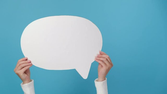 Close up cropped shot of female hands holding an empty white speech bubble, shaking left and right, isolated on blue studio background with copy space for advertisement. Dialog concept