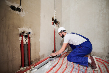 Wide angle view on young worker standing on his knees in blue overalls and helmet installing...