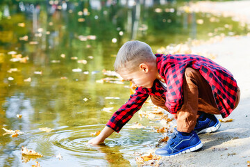 A little boy plays in the water near a natural lake with fallen leaves and sun glare. Sitting on a sandy beach, he pulls something out of the water with his hand.