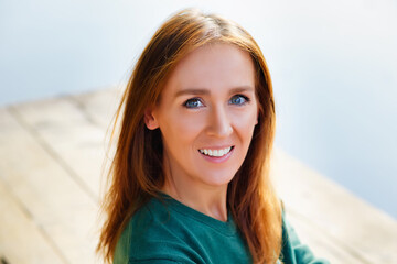 A large portrait. A young, positive, smiling woman sits on the wooden railing of the pier, a blurry lake landscape in the background.