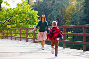 Obraz premium A beautiful woman and a boy on a wooden bridge. Selective focus. A son in a red checked shirt runs to meet his mother.