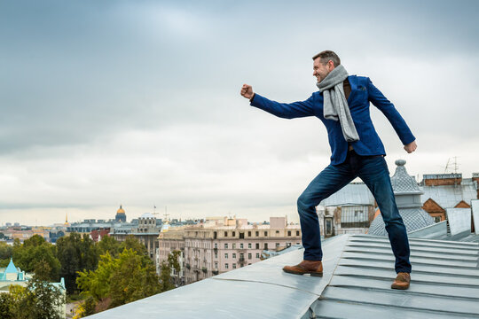 Happy stylish young man in jeans, a blue jacket and a scarf stands on the edge of the roof of a building in a European city. The person shows a hand gesture with a clenched fist. St Petersburg, Russia