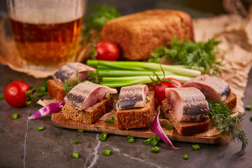Slices of salted herring on black bread, light beer and fresh vegetables on a wooden kitchen board...