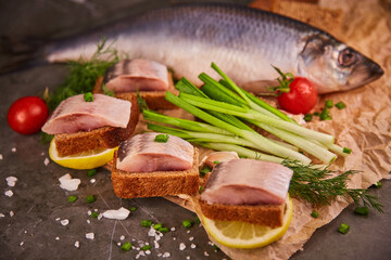Slices of salted herring on black bread, lemon, fresh vegetables and green onions on a wooden kitchen board on paper. Delicious snack at the bar