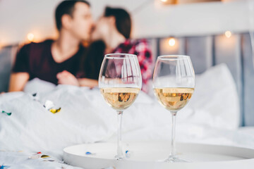Glasses of white wine in the foreground and out of focus happy young man and woman kiss on bed in light hotel room closeup.