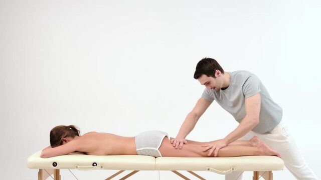 Male masseur with strong hands massaging lower part of leg to young woman lying on massage table in spa salon. Professional physiotherapist performing foot and lower leg massage in white background.