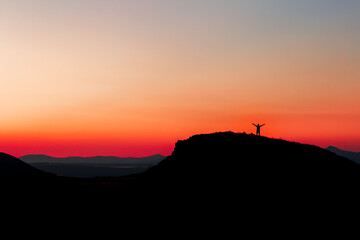 Red sunset with mountain silhouette. A man on mountain in sunset. Traveler on rock waving hands.