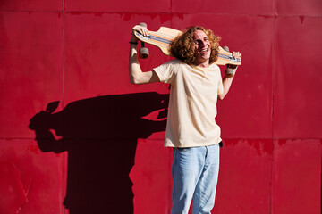 young man with curly hair looking at the camera with his skateboard on his shoulders and a red background
