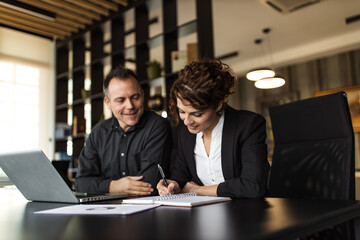 Portrait of a business woman writing in note pad and business man looking while sitting at desk with laptop.