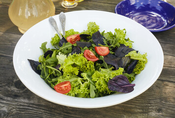 Fresh salad mix of arugula leaves, basil, lambs lettuce and tomatoes. Salad bowl, healthy food. Composition in a white plate on an old wooden table