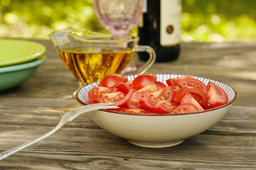 Sliced tomatoes in a bowl are ready to eat. Composition in a fresh authentic atmosphere. Oil, plates and wine in the background