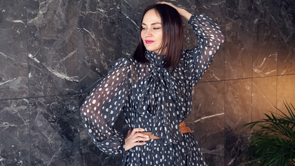 Short haired brunette in grey polka dot dress with brown belt poses for camera smiling near marble wall with white patterns in studio