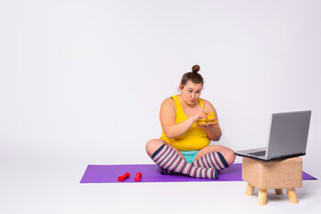 Large sporty woman working out at home on the yoga mat in front of the open laptop repeats the instructions of a professional fitness trainer and eat biscuit. White background.