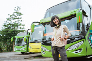 An Asian woman is standing in a backpack and headphones smiling against the background of the bus