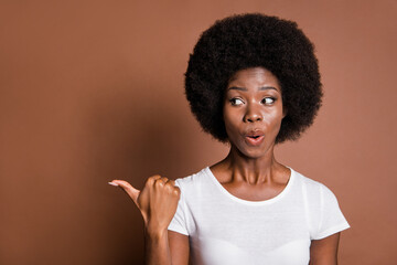 Photo portrait of girl pointing thumb on blank space advising curious isolated on brown color...